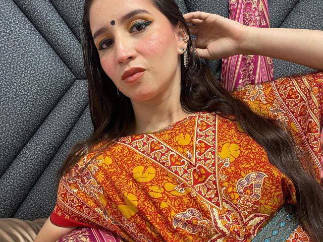 JalimaMona with long brown hair, pink blush, yellow eyeshadow, small black dot on forehead, wearing an orange and red patterned top, lying back with hand in hair.