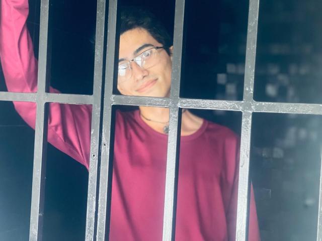 AnryuOswald with glasses, wearing a red shirt, standing behind black metal bars, arm raised, looking through the bars with a slight smile.