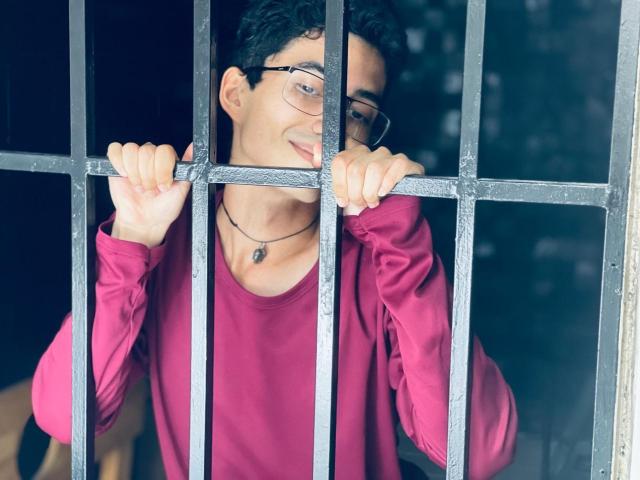AnryuOswald with glasses, black curly hair, pink shirt, and necklace, gripping metal bars with both hands, smiling slightly, standing behind bars.