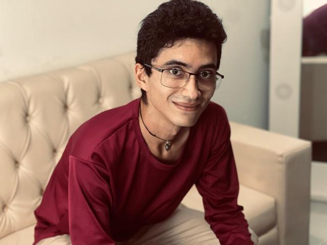AnryuOswald, a cute dark-haired dude with glasses, wearing a red shirt and black necklace, leaning on a beige tufted sofa, smiling softly at the camera.