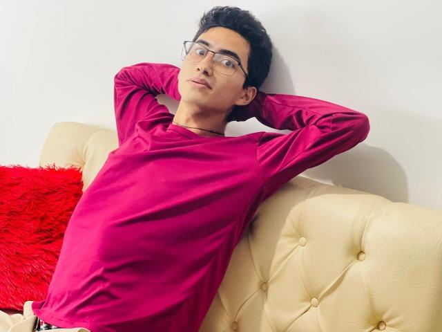 AnryuOswald, with glasses and dark hair, wearing a bright pink shirt, leans back on a cream sofa, arms behind head, red fuzzy pillow to the left.