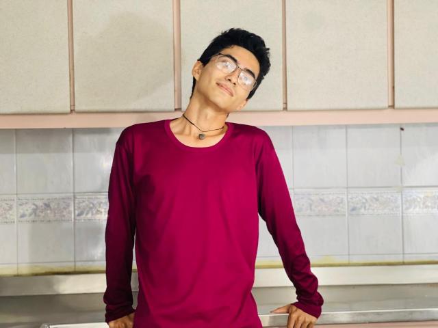 AnryuOswald with short black hair, glasses, and a red long-sleeve shirt, leaning back against a kitchen counter, wearing a black necklace.