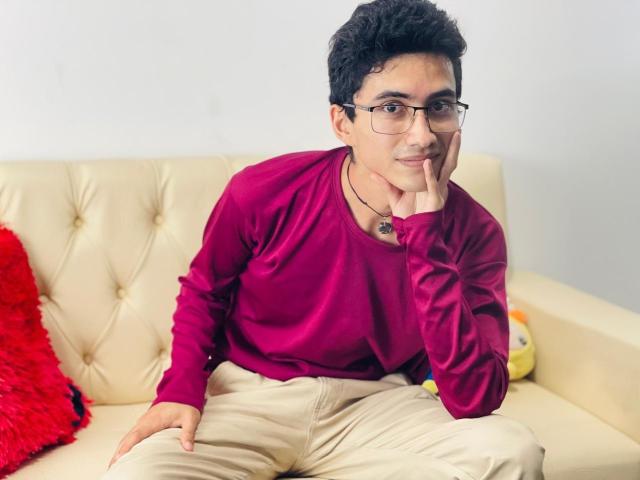 AnryuOswald, short curly black hair, glasses, red shirt, beige pants, hand on cheek, red fuzzy pillow, beige tufted sofa, small stuffed toy on right.