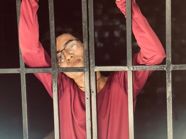 AnryuOswald with glasses, arms through black metal bars, wearing a red long-sleeve shirt, looking up with a slightly open mouth, short curly hair, medium skin tone, serious expression.