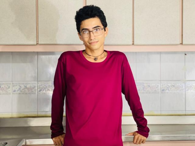 AnryuOswald, short black-haired dude, wearing glasses, red long-sleeve shirt, necklace with small pendant, standing in kitchen, hands on counter, slight smile, casual, cute.