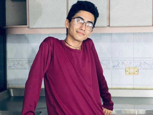AnryuOswald with curly black hair, glasses, and a red long-sleeve shirt, standing in a kitchen, smiling, hands on a stainless-steel counter.