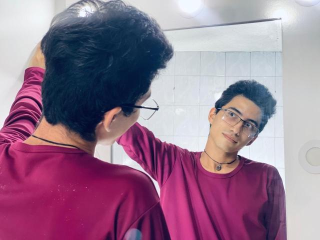 AnryuOswald with curly black hair and glasses, wearing a red shirt, stands in front of a mirror, touching his head, looking at his reflection with a slight smile.