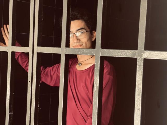 AnryuOswald, with glasses and a red shirt, smiling through metal bars, has a necklace and is touching the bars with his right hand.