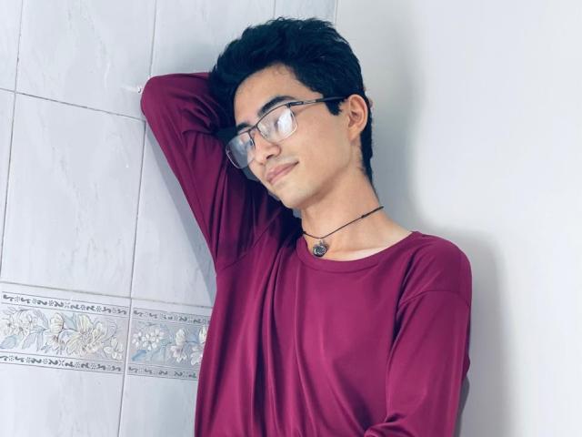 AnryuOswald, nerdy dude with glasses, wearing a maroon shirt, leaning against a wall, arm raised, necklace with a small pendant, smiling slightly, short black hair.