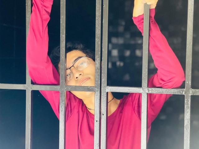 AnryuOswald with glasses, arms raised, wearing a pink shirt, standing behind black metal bars, looking through them with a slightly tired expression.
