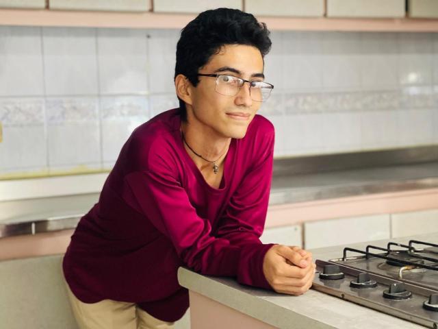 AnryuOswald with curly black hair, glasses, and a red shirt leans on a kitchen counter, smiling slightly, wearing beige pants and a necklace with a small cross.