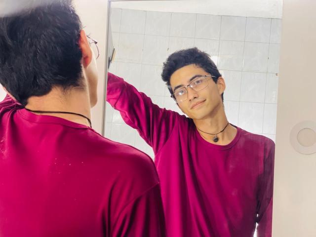 AnryuOswald with dark curly hair, glasses, and a red shirt, stands in front of a mirror, touching it with his right hand, wearing a black necklace.