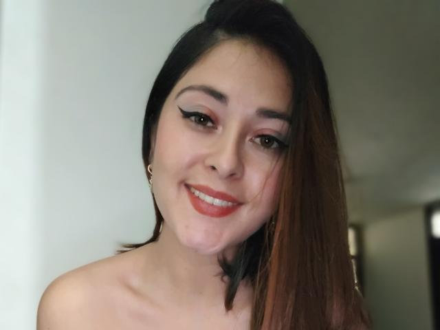 LisbethMooree has a cute smile with red lipstick, dark eyeliner, and brown eyes. She's topless with straight brown hair, wearing small gold earrings. Her tits are not visible.