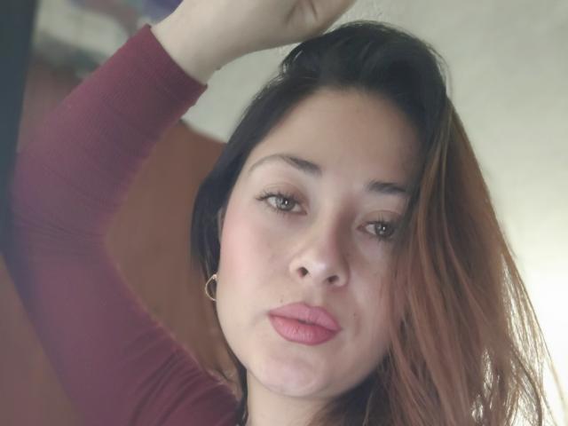 LisbethMooree with puffy pink lips, light brown eyes, and dark brown hair, wearing a red long-sleeve top, raising her right arm, showing a gold hoop earring.