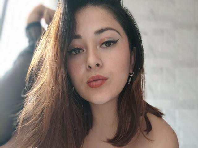 LisbethMooree has medium brown hair, fair skin, black eyeliner wings, and orange lipstick. She's looking at the camera with a pouty expression, wearing silver earrings.