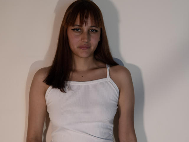 LiaMonroelu has straight brown hair, a white ribbed tank top, and a nose ring. Her tits are small and she's looking straight at the camera with a neutral expression.