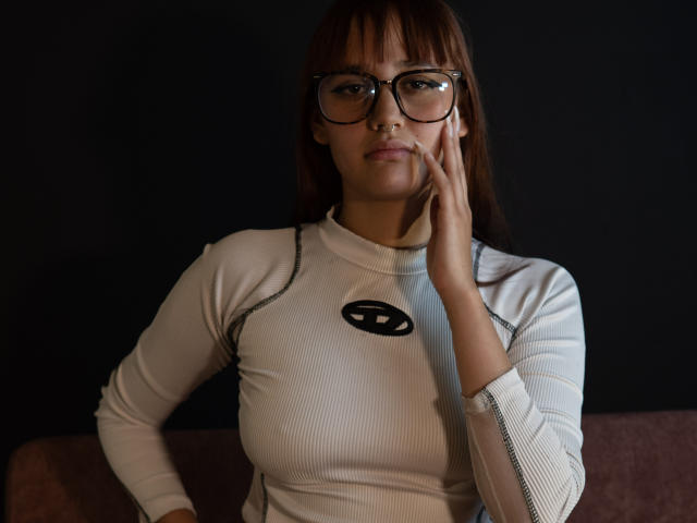 LiaMonroelu with glasses, white ribbed top, black background, hand on cheek, brown hair, small tits, dark eyes, serious expression, dark skin, soft light, casual pose.