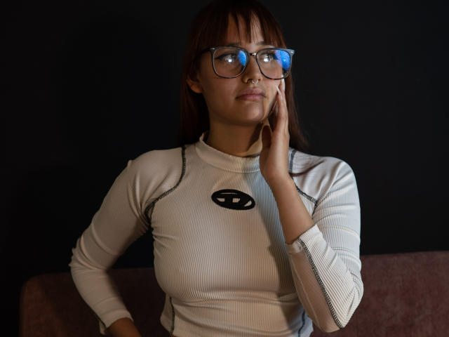 LiaMonroelu with glasses, white ribbed long-sleeve top, black nose ring, hand on cheek, brown hair, dark background, looking slightly to the side, small tits, serious expression.