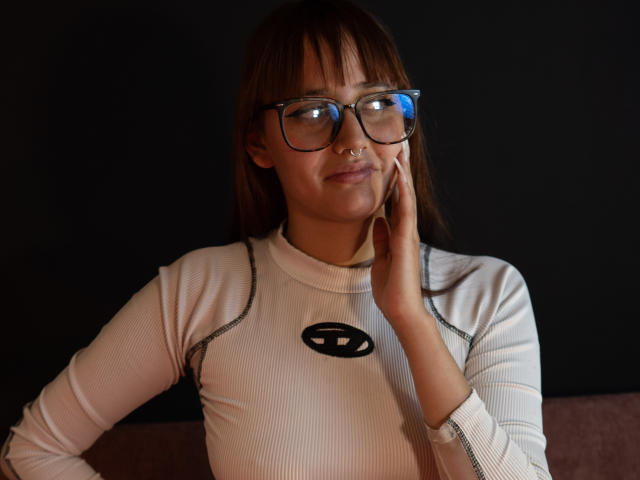 LiaMonroelu with brown hair, glasses, nose ring, white ribbed turtleneck, black logo, touching her cheek, slightly smiling, dark background. Boobs not visible, focus on upper body.