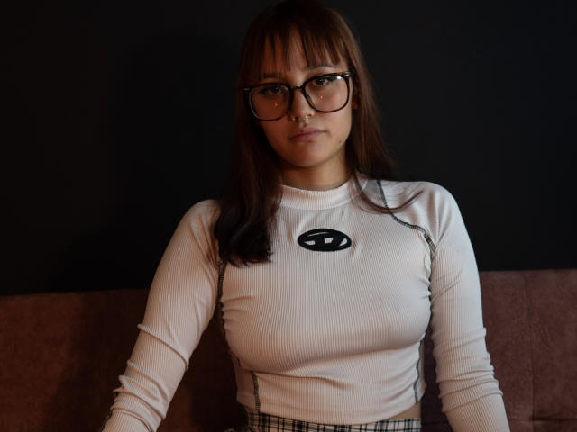 LiaMonroelu with glasses, white ribbed shirt, small black eye logo, and plaid pants, sitting on a brown couch, looking serious, brown hair, bangs, medium tits.