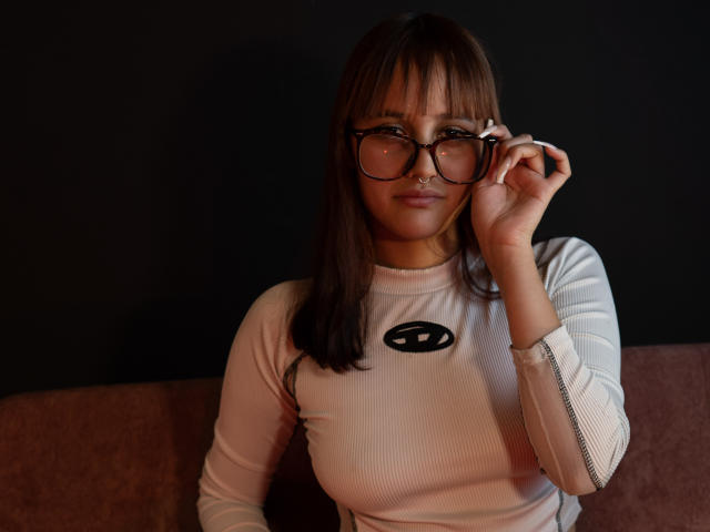 LiaMonroelu with brown hair, wearing glasses, white ribbed long-sleeve shirt, adjusting glasses with right hand, small nose ring, sitting on brown couch, dark background.