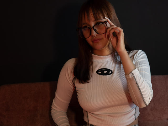 LiaMonroelu with glasses, adjusting them with her right hand, wearing a white long-sleeve shirt with a black logo, sitting on a pink couch, looking slightly down.