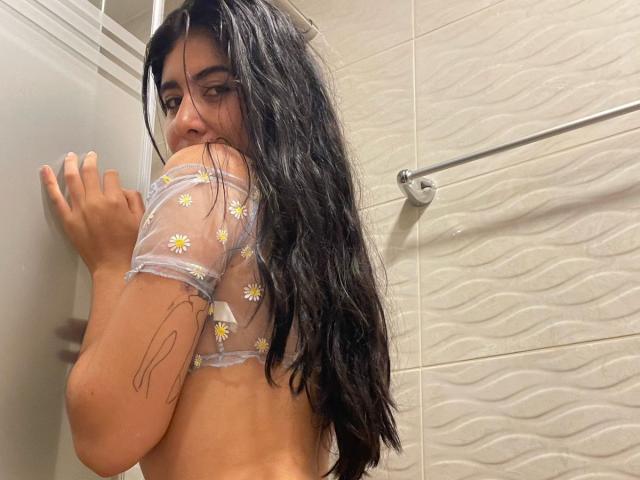 MiluSky, avec ses cheveux noirs et mouillés, porte un haut transparent à motifs de marguerites, laissant apparaître son dos nu et ses petites seins. Elle regarde par-dessus son épaule, une main sur un verre, et a un tatouage sur le bras..