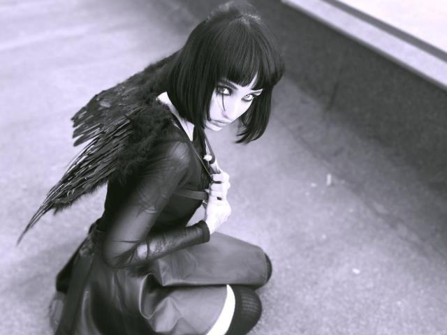 HollyVex a des ailes sombres, une coupe de cheveux courte, et est agenouillée, vêtue de noir de style gothique, avec des bas hauts, les yeux levés avec intensité, tenant sa poitrine. Photo en noir et blanc..