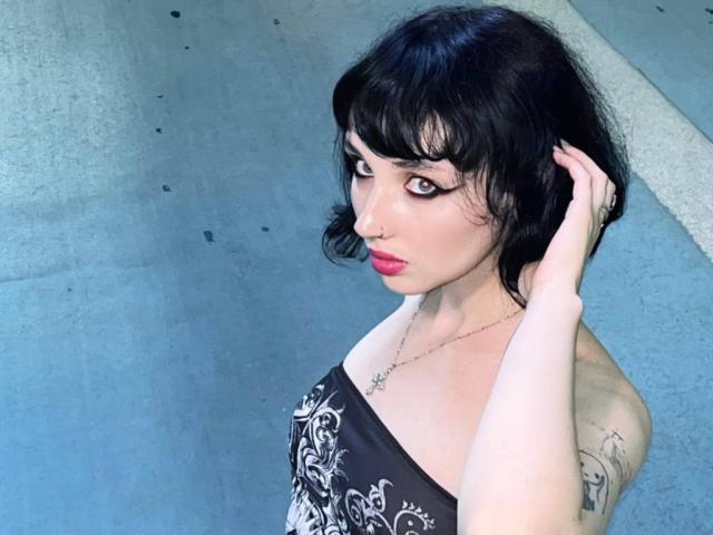 HollyVex with black hair, bangs, and red lipstick, wearing a strapless black dress with white patterns, touching her hair, showing a tattoo on her right arm.