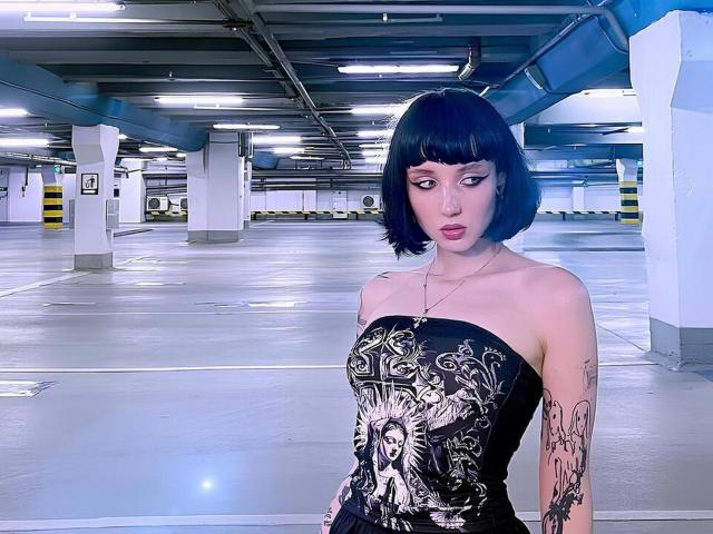 HollyVex has black bob, wearing strapless black and gold dress with sun face, tattoos on arm, standing in empty parking garage, looking serious, pale skin, blue eyes, pink lips.