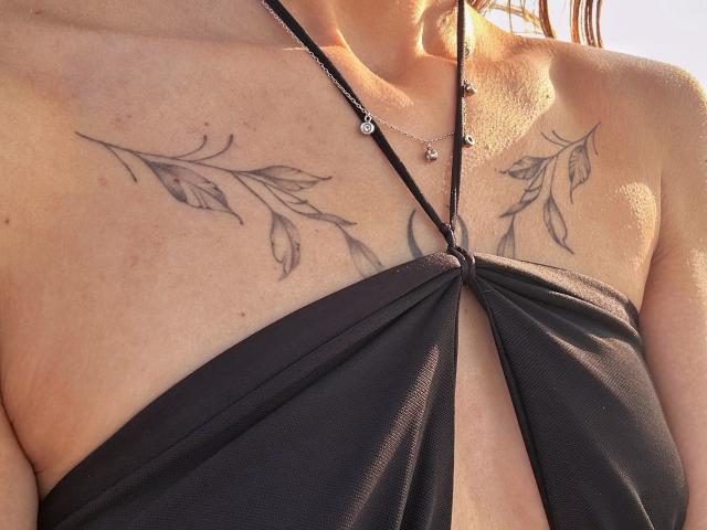 Karishs Brust mit schwarzen Blatt-Tattoos, trägt ein schwarzes Halstuch-Top, das tiefe Ausschnitte zeigt, eine Halskette mit kleinen Anhängern, sonnengebräunte Haut, braunes Haar und einen leichten Teint..