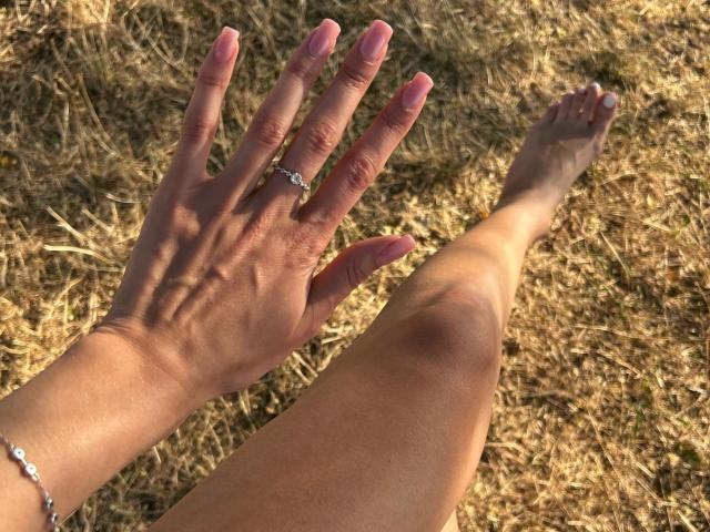 La mano de Karish, con uñas rosas y un anillo de plata, y que lleva una pulsera, descansa sobre la hierba seca. La pierna desnuda de Karish, con un ligero moretón, está extendida junto a su mano..