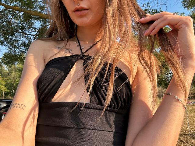 Karish with long blonde hair, black halter top, small tattoos, and a silver bracelet, holding her hair, showing her bare shoulders and chest. Sunlight casts shadows on her.