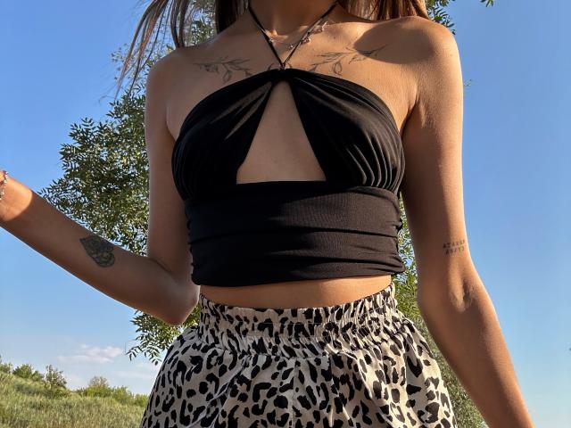 Karish, avec de petites seins, portant un haut noir à enfiler et une jupe à motifs léopard, avec des tatouages sur son bras droit et son épaule gauche, debout à l'extérieur, au soleil..