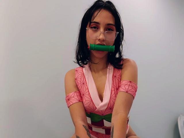 SofiaaMiiller with black hair, glasses, pink geometric top, green belt, and green dildo in mouth, sitting, arms crossed, tattoo on right wrist.