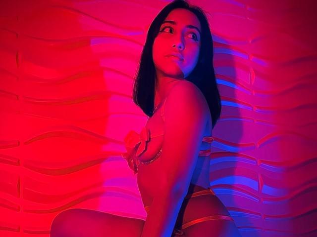 SofiaaMiiller with dark hair, red and blue lighting, covering her tits with a hand, wearing strappy lingerie, sitting against a wavy wall, looking up with a serious expression.