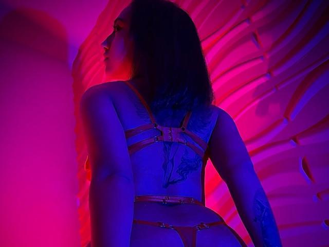 SofiaaMiiller in pink and blue neon, wearing a strappy red harness, showing her tattooed back and butt, with her head turned to the side, looking seductive.
