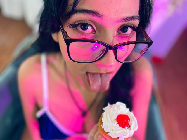 SofiaaMiiller with black glasses, sticking out tongue, holding a cupcake with a cherry, pink and purple lighting, dark hair, and a blue top. Boobs visible under top.