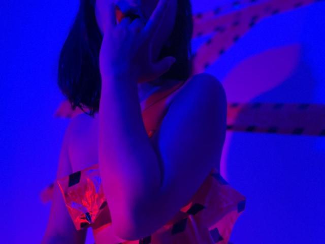 SofiaaMiiller with purple and pink neon lighting, covering her mouth with one hand, wearing a red floral dress, her bare shoulder and upper chest are visible.