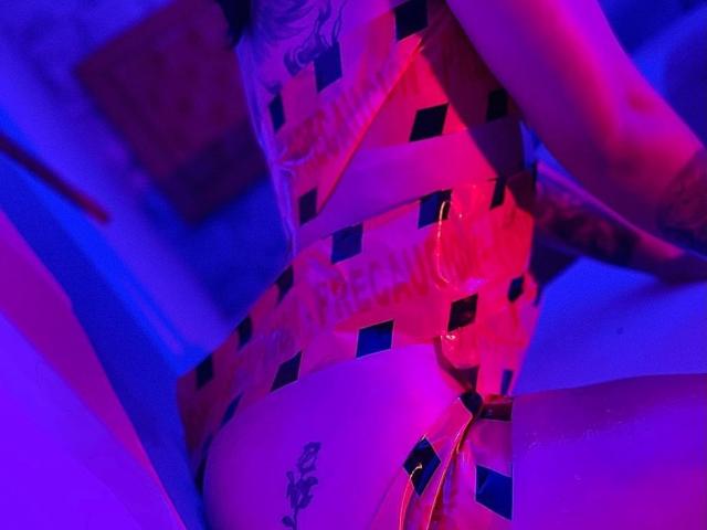 SofiaaMiiller in a red and black checkered latex bodysuit, hands on her chest, purple and pink neon lights, tattoo on her right thigh, tattoo on her left arm.