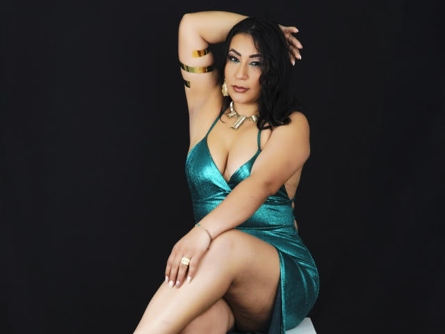 EmaFoxy in a shiny green dress, showing off her boobs and ass with a raised arm, wearing gold bracelets and a necklace, sitting against a black background.