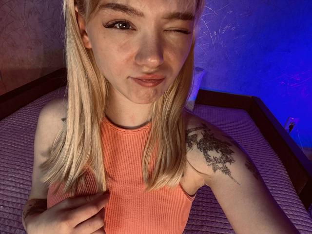 EnchantingEyes, blonde, winking, in a ribbed orange tank top, showing a tattoo on her right shoulder, holding her top with her left hand, blue light in background.