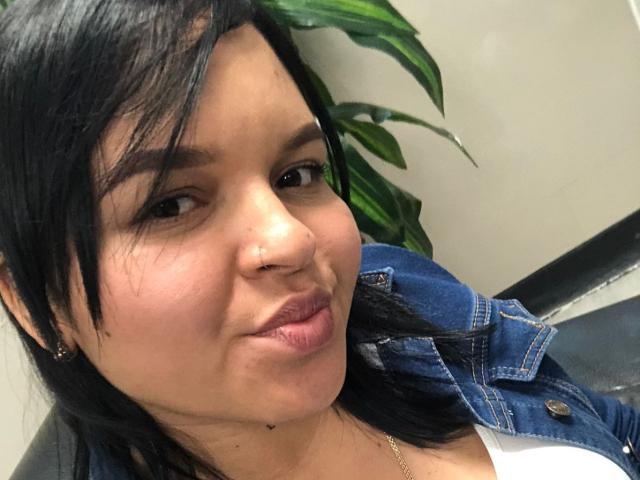 YeriBellani has black hair, a nose piercing, and is wearing a denim jacket with a white shirt, pouting with pink lipstick, and a gold necklace, looking at the camera.