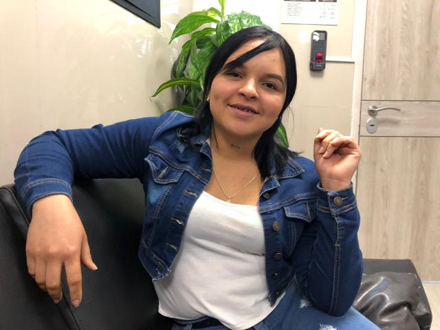 YeriBellani, a chubby woman with black hair, wearing a denim jacket and white top, sitting on a black couch, smiling slightly, holding a cigarette.