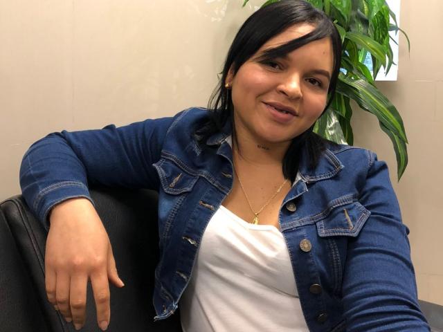 YeriBellani with black hair, wearing a blue denim jacket and white top, sitting, arms resting, slight smile, small gold necklace, green plant in background.
