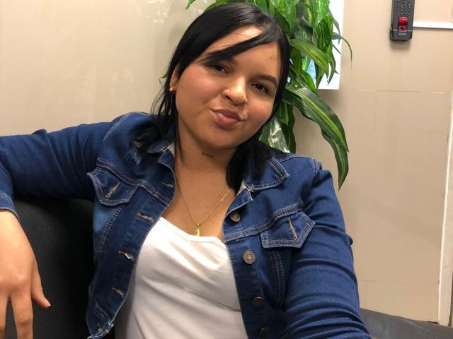 YeriBellani with black hair, pouty lips, wearing a blue denim jacket over a white tank top, small gold necklace, and gold earrings, leaning on a black chair.
