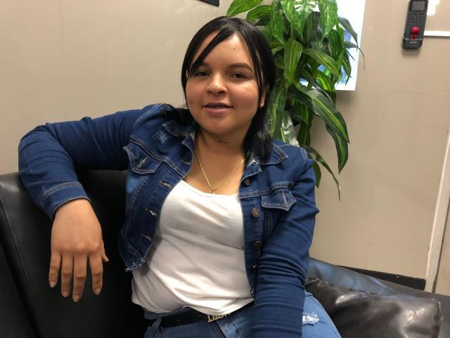 YeriBellani with black hair, wearing a blue denim jacket and white top, sits on a black leather chair, smiling, hands relaxed, showing a small tattoo under her chin.