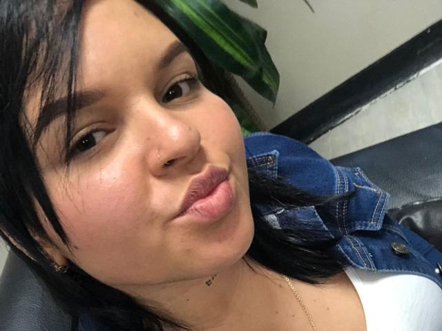 YeriBellani with black hair, brown eyes, nose ring, and pouty lips, wearing a blue denim jacket over a white top, sitting on a black leather chair.