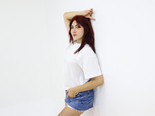 SpencerLe met rood haar, een wit T-shirt en blauwe denim shorts, arm omhoog, hand in de zak, staand tegen een witte muur, lichtelijk opzij kijkend, met een zelfverzekerde uitdrukking..