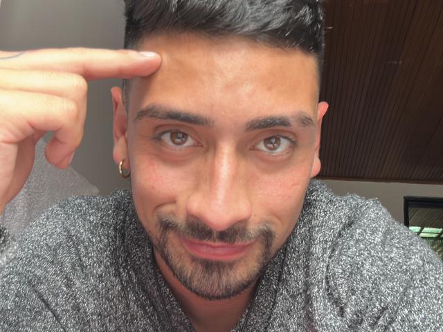 ApoBe has a cute, handsome face with brown eyes, short black hair, and a small beard. He's pointing at his forehead with his right finger, wearing a gray sweater.